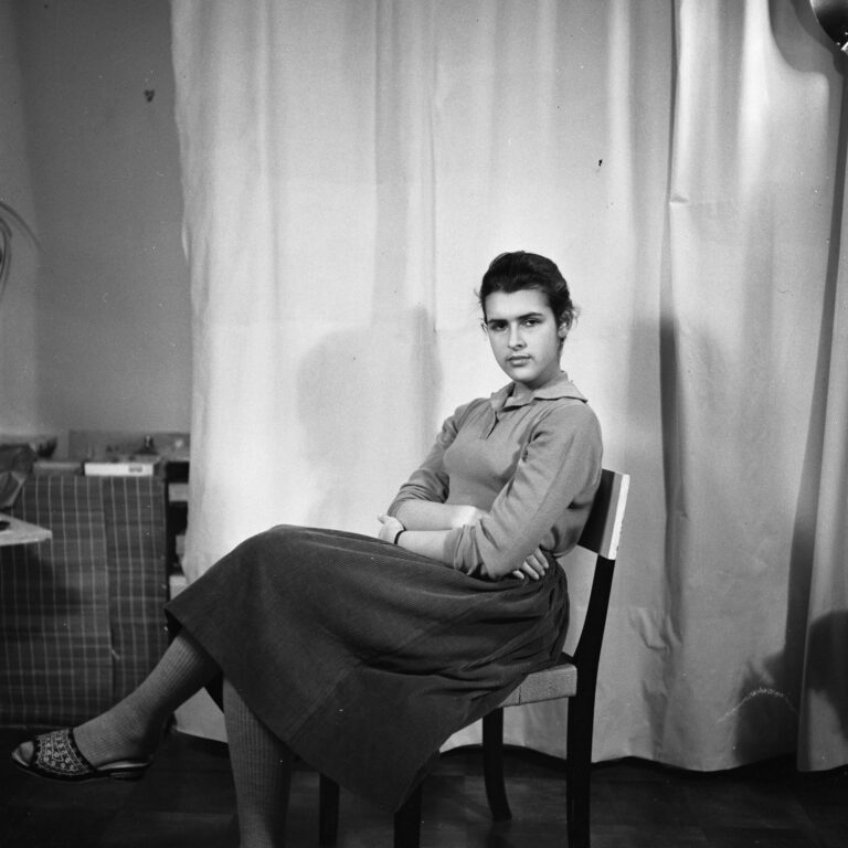 Svartvitt porträttfotografi av Vera som sitter på en stol i ateljén med benen och armarna i kors. Tittar in i kameran med bestämd blick.