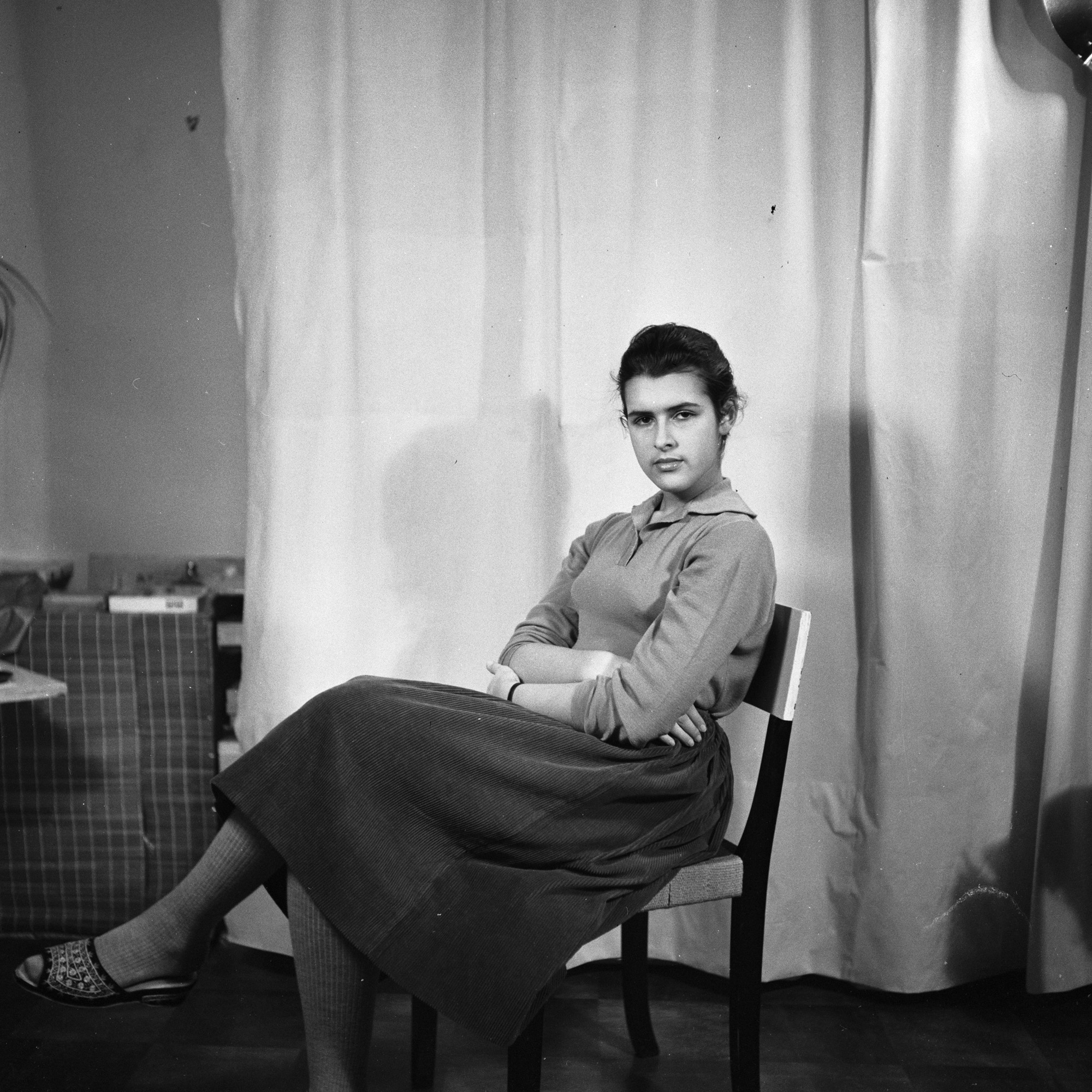 Svartvitt porträttfotografi av Vera som sitter på en stol i ateljén med benen och armarna i kors. Tittar in i kameran med bestämd blick.